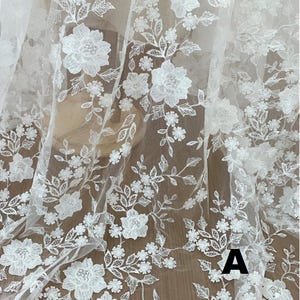 May include: White floral lace fabric with detailed embroidered flowers and leaves. The delicate design features various sizes of blossoms and foliage, creating a textured, elegant pattern. The fabric is semi-transparent, suitable for bridal or decorative projects.