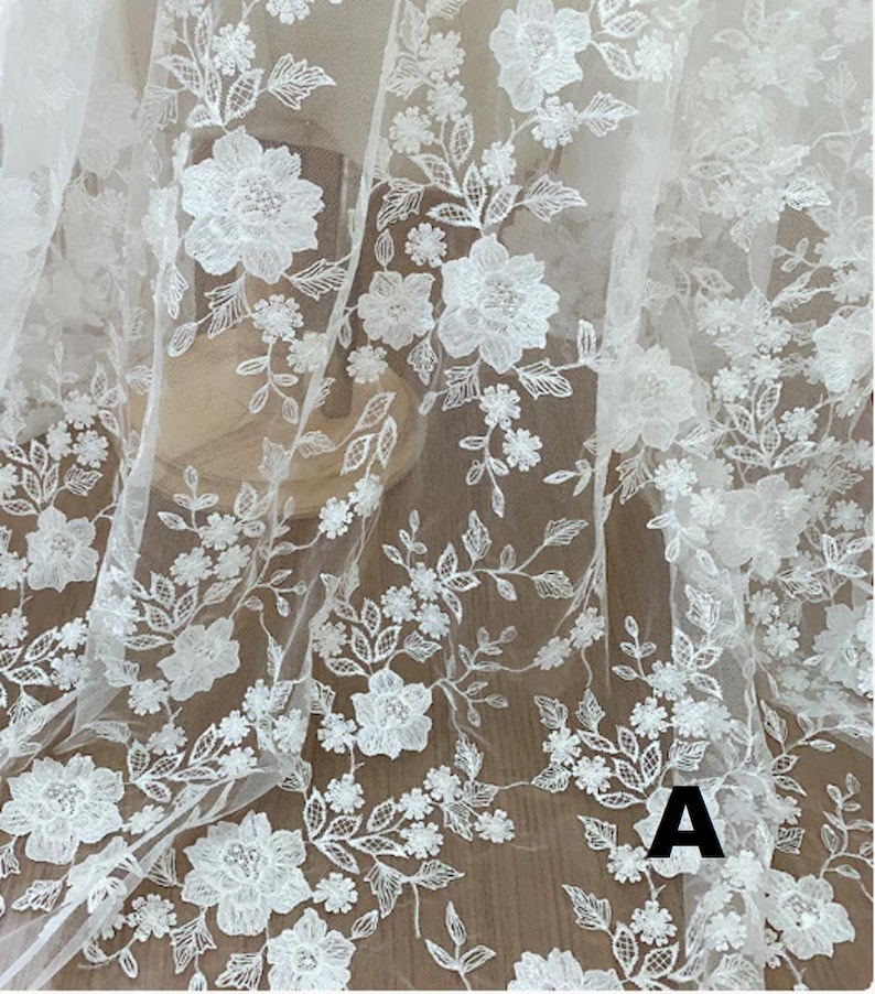 May include: White floral lace fabric with detailed embroidered flowers and leaves. The delicate design features various sizes of blossoms and foliage, creating a textured, elegant pattern. The fabric is semi-transparent, suitable for bridal or decorative projects.