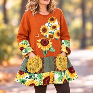 May include: A long-sleeve tunic featuring a burnt orange top section with a sunflower print. The lower portion is a patchwork of green, brown, and plaid fabrics with two yellow crocheted pockets and a sunflower-patterned ruffled hem.