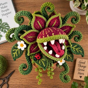 May include: A crocheted carnivorous plant in a terracotta pot featuring deep red and green leaves, curling vines, white teeth, a pink tongue, and a small red ladybug. The plant is surrounded by green yarn, scissors, and signs.