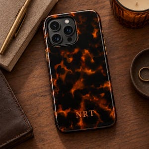 Puede incluir: Una funda rectangular para smartphone con un patrón brillante de carey en tonos negro y ámbar. La funda tiene un recorte para cámara de triple lente en la parte superior izquierda y las letras NRT impresas en fuente dorada en el centro inferior.