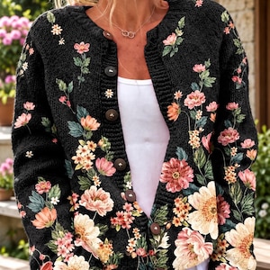 May include: A black knit cardigan featuring a floral pattern with pink, orange, and yellow flowers and green leaves. The garment has a round neckline, long sleeves, and a front button closure with dark brown buttons over a white undershirt.