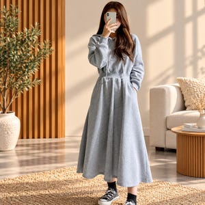 Puede incluir: Un vestido largo gris claro con capucha, mangas largas, cintura entallada y falda acampanada. El vestido incluye una capucha con cordón, bolsillos laterales y una textura suave. Se combina con calcetines negros y zapatillas deportivas.