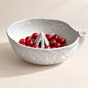 May include: A white ceramic citrus juicer bowl with a textured, wavy exterior surface and a central reamer cone. The bowl is filled with numerous small, round, bright red cranberries, and features a small pouring spout on the right side.
