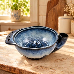May include: A ceramic yarn bowl featuring a deep blue and white mottled glaze. The bowl has a smooth, rounded shape with a side opening for yarn, a small central dome with holes, and a flat handle extension on the left side with three small holes.