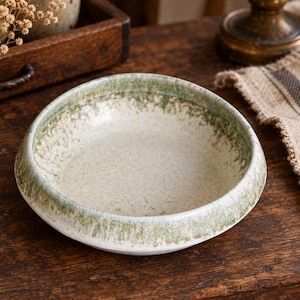 Puede incluir: Un cuenco de cerámica redondo y poco profundo con una base de color blanco roto moteado y un degradado de esmalte verde musgo texturizado alrededor del borde grueso y redondeado. La superficie interior tiene un aspecto sutilmente manchado, mientras que el borde exterior muestra una transición clara desde el centro claro hacia el pigmento verde tierra más oscuro.