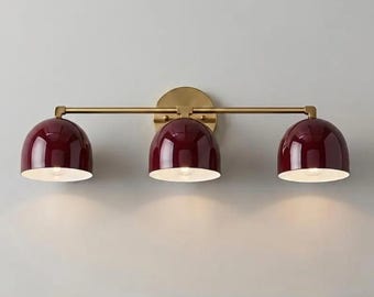 Puede incluir: Un aplique de pared con una barra horizontal de latón unida a una placa circular de latón. Tres pantallas brillantes en forma de cúpula de color burdeos intenso están espaciadas a lo largo de la barra, cada una con interior blanco.