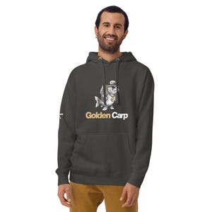 May include: A dark gray hooded sweatshirt featuring a graphic of a fish wearing a baseball cap, sunglasses, and a gold chain. Below the fish, the text reads Golden Carp in gold and white lettering. The hoodie includes a front kangaroo pocket.