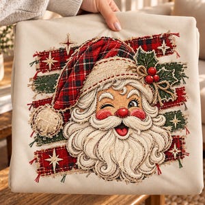 May include: A beige sweatshirt featuring a central graphic of a winking Santa Claus wearing a red and black plaid hat. The design includes frayed fabric strips in red and green, holly berries, and embroidered star accents on a light background.