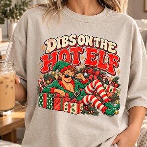 Peut inclure: Un t-shirt beige à manches courtes avec un motif représentant un elfe portant des lunettes de soleil et une tenue verte, allongé parmi des cadeaux de Noël. Le texte DIBS ON THE HOT ELF est écrit en grandes lettres rouges et crème.