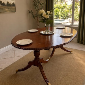 Puede incluir: Una mesa de comedor ovalada de madera con acabado en marrón oscuro pulido, con un borde tallado en forma de cuerda, una base de pedestal central y cuatro patas abiertas que terminan en garras de metal dorado con ruedas, con cuatro platos.