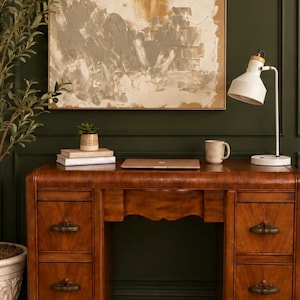 May include: A wooden desk with four drawers featuring dark metal pulls, a closed laptop, a small potted succulent on two stacked books, a ceramic mug, and a white desk lamp with a wooden accent, set against a dark green wall.