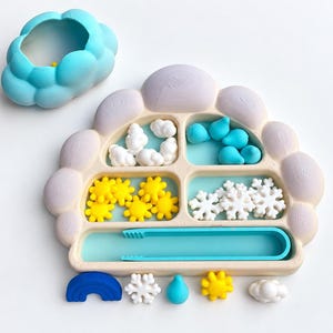 Puede incluir: Una bandeja dividida con forma de nube en color beige claro con un cuenco de nube azul a juego. La bandeja contiene pequeñas figuras blancas de nubes, gotas de lluvia azules, soles amarillos y copos de nieve blancos, junto a unas pinzas azules.