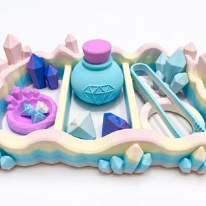 Puede incluir: Una bandeja rectangular con borde degradado en tonos pastel y formas de cristal esculpidas. Contiene una botella azul claro con un icono de diamante, un cuenco morado con cristales pequeños, gemas con forma de diamante y unas pinzas azules.
