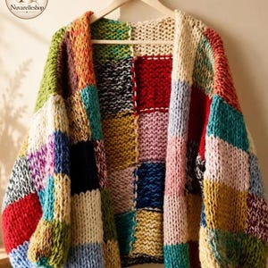 May include: A chunky knit cardigan featuring a patchwork design of large squares in various colors including red, yellow, blue, green, pink, and cream. The garment has long sleeves, a loose fit, and is displayed on a wooden hanger with a logo.
