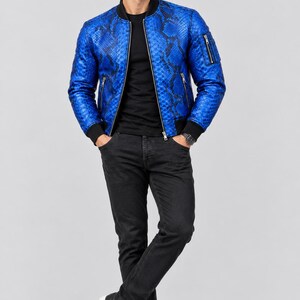 Könnte beinhalten: Eine leuchtend blaue Bomberjacke mit schwarzem Schlangenledermuster, schwarzem Rippstrickkragen, Bündchen und Saum sowie silberfarbenen Metallreißverschlüssen an der Vorderseite und der Ärmeltasche, getragen über einem schwarzen T-Shirt und Jeans.