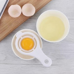 May include: A white plastic egg separator tool resting on a small white bowl, containing a bright yellow egg yolk. A separate white bowl holds the clear egg white, and two empty eggshell halves are positioned nearby on a wooden surface.
