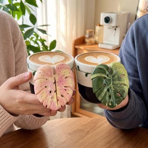 May include: Two ceramic mugs filled with latte art coffee. One mug features a pink and yellow variegated monstera leaf handle, while the other has a deep green monstera leaf handle. Both mugs have a white upper section and a dark base.