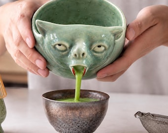 以下が含まれることがあります： 尖った耳と注ぎ口のような口を持つ生き物の形をした淡い緑色の陶器のボウル。その口から鮮やかな緑色の液体が、下に置かれた暗い質感のメタリック仕上げの陶器のボウルへと注がれている様子。