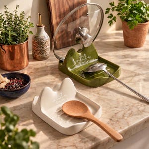 May include: Two ceramic spoon rests, one olive green and one cream, each featuring a central indentation for utensils and a back slot for a pot lid. The green rest holds a metal lid and spoon, while the cream rest holds a wooden spoon.