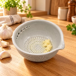 May include: A light gray ceramic garlic grater bowl featuring a textured, flower-shaped grinding surface at the bottom and a series of small holes along the inner rim for herb stripping, containing a small amount of freshly grated garlic.