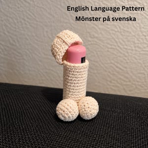 May include: A cream-colored crochet cover for a cylindrical object, featuring a phallic shape with a separate cap piece. The text 'English Language Pattern' and 'Mönster på svenska' is visible at the top of the image, with a pink object inside.