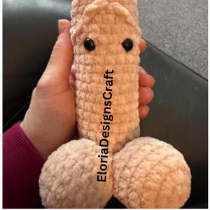 May include: A beige, phallic-shaped crochet plush toy made of soft, textured yarn. It features two round base sections, a cylindrical body, and two small, black, circular plastic eyes. The text EloriaDesignsCraft is printed vertically on the front.