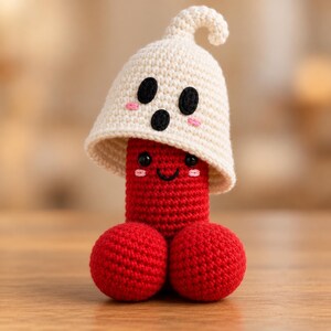 May include: A handcrafted crochet plush toy featuring a bright red cylindrical body with two round red spheres at the base. The figure has a smiling face with black eyes and pink cheeks, topped with a cream-colored ghost hat with black eyes.
