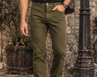 May include: A black short-sleeved t-shirt, olive green denim jeans, dark brown leather boots, a brown leather shoulder bag, a silver chain necklace, and dark-tinted sunglasses. The outfit is worn with a silver wristwatch on the left wrist.