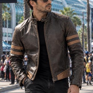 Puede incluir: Una chaqueta de cuero marrón con cuello alzado, cremallera frontal y dos rayas horizontales de color beige en cada manga. La chaqueta se lleva sobre una camiseta negra y pantalones cargo negros, junto con gafas de sol oscuras.