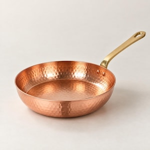 Può includere: Una padella rotonda in rame martellato con un manico in metallo lucido color oro. La padella presenta una superficie testurizzata distinta creata da piccole rientranze uniformi sulle pareti esterne ed interne, fissata con due rivetti.