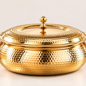 May include: A round, gold-coloured serving dish with a hammered texture finish. It features two side handles, a matching lid with a central knob, and a smooth, reflective surface. The design is consistent across the body and the lid.