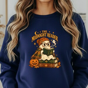 May include: A navy blue crewneck sweatshirt featuring a graphic of a ghost wearing a beanie and glasses, reading a book while sitting on a stack of books next to a pumpkin. The text above the ghost reads THE MIDNIGHT READER in yellow.