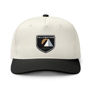 May include: A cream-colored baseball cap with a black brim and a black button on top. The front features a shield-shaped patch with a black background, a gold sun, a white mountain graphic, and the text YELLOWSTONE EST 1872 NATIONAL PARK.