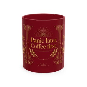 May include: A deep red ceramic mug featuring gold-colored decorative line art and the text "Panic later. Coffee first." printed in a serif font. The design includes symmetrical geometric borders and stylized wheat or coffee bean motifs.
