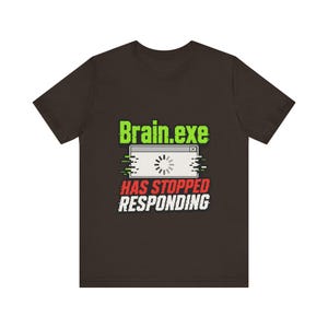 Peut inclure: Un t-shirt à manches courtes marron foncé avec un imprimé graphique affichant le texte "Brain.exe" en vert, une icône de fenêtre de chargement blanche et le texte "HAS STOPPED RESPONDING" en rouge et blanc avec un effet de glitch numérique.