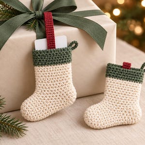 May include: Two small, cream-coloured crocheted stockings with dark green cuffs and loops. The larger stocking features a red fabric tab and holds a white rectangular card. The items are displayed against a gift box with a green ribbon.