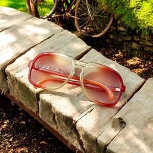 May include: Retro-style sunglasses with a gradient lens, transitioning from red to yellow. The frames are a combination of red and clear plastic, with a classic square shape. The sunglasses are resting on a weathered wooden surface.