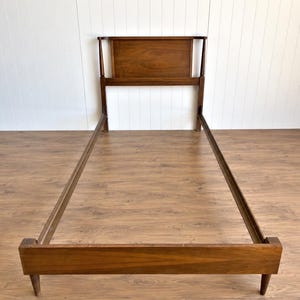 May include: A single bed frame constructed from dark brown wood, featuring a rectangular headboard with a smooth panel, two vertical side supports, long side rails, and a low footboard supported by four tapered legs in a simple design.
