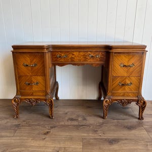 May include: A wooden desk featuring a polished brown finish with a prominent wood grain pattern. The desk includes a central drawer flanked by two columns of two drawers each, all equipped with ornate metal handles and supported by carved legs.