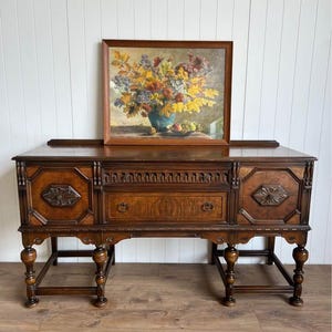 May include: A dark brown wooden sideboard featuring two side cabinets with carved floral medallions, a central drawer with two metal ring pulls, and turned legs. A framed oil painting of a colorful floral bouquet sits on top.