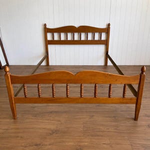 May include: A wooden bed frame featuring a headboard and footboard with a medium brown finish. Both the headboard and footboard have a curved top rail and a series of vertical, turned spindle posts. The frame includes side rails connecting the two.
