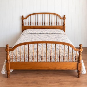 May include: A wooden bed frame with a spindle headboard and footboard, featuring a warm brown finish. The bed is dressed in a cream-colored quilt with a repeating blue floral pattern, covering the mattress and extending over the footboard.