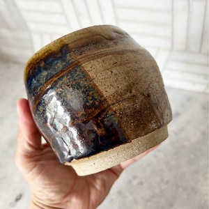 May include: A small, round ceramic cup with a textured, unglazed beige base and a glossy, dark brown and deep blue glazed upper section. The surface features visible clay imperfections and a distinct transition between the matte and shiny finishes.