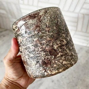 May include: A cylindrical ceramic vessel featuring a textured, mottled surface with a dark brown, black, and cream-colored finish. The exterior has a cratered, uneven appearance with a glossy sheen, showcasing a complex, organic pattern throughout.