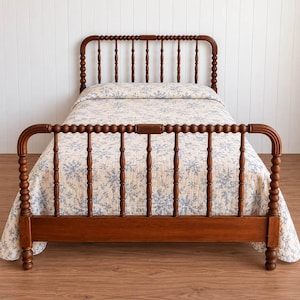 May include: A wooden bed frame with a spindle design featuring a tall headboard and a lower footboard. The bed is dressed in a cream-colored quilt with a repeating blue floral pattern. The frame has a warm brown finish and rounded posts.