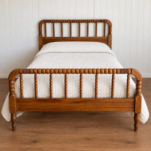 May include: A wooden bed frame featuring a headboard and footboard with a spindle design composed of rounded, bead-like segments. The bed is dressed in a white quilted coverlet, showcasing a warm brown wood finish throughout the frame.