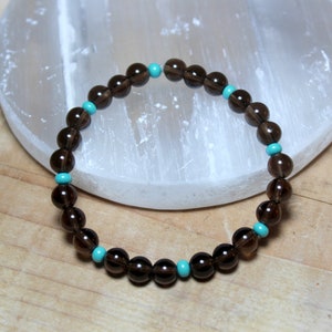 May include: A beaded bracelet featuring round, translucent dark brown smoky quartz beads alternating with small, vibrant turquoise blue accent beads arranged in a circular pattern to form a continuous loop of polished gemstone spheres.
