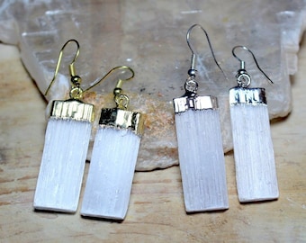 Puede incluir: Tres pares de pendientes de cristal de selenita con detalles dorados y plateados. Los pendientes son de forma rectangular y tienen un aspecto blanco opaco.