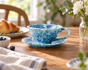 Könnte beinhalten: Eine Keramiktasse mit passender Untertasse, die einen gewellten Rand und eine gesprenkelte Aquarell-Glasur in Blau-, Grün- und Weißtönen aufweist. Die Tasse hat einen geschwungenen Henkel und steht mittig auf der Untertasse mit glänzender Oberfläche.